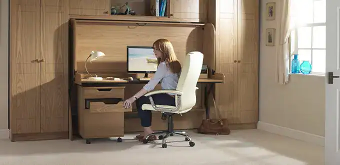 A girl studying at a wooden desk with a computer in a cosy, well-lit room with wooden wall panels and a window, illustrating a comfortable and functional study environment for students.